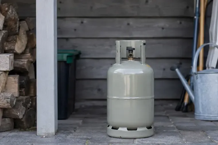 Een metalen gasfles staat rechtop op een koele, beschutte plek naast een houten schuur