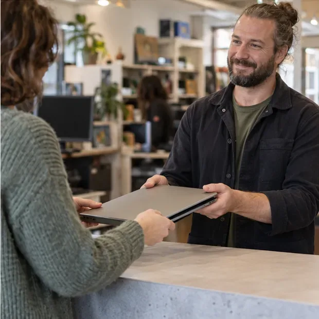 Een bezoeker geeft een gesloten laptop aan een medewerker achter een neutrale innamebalie.