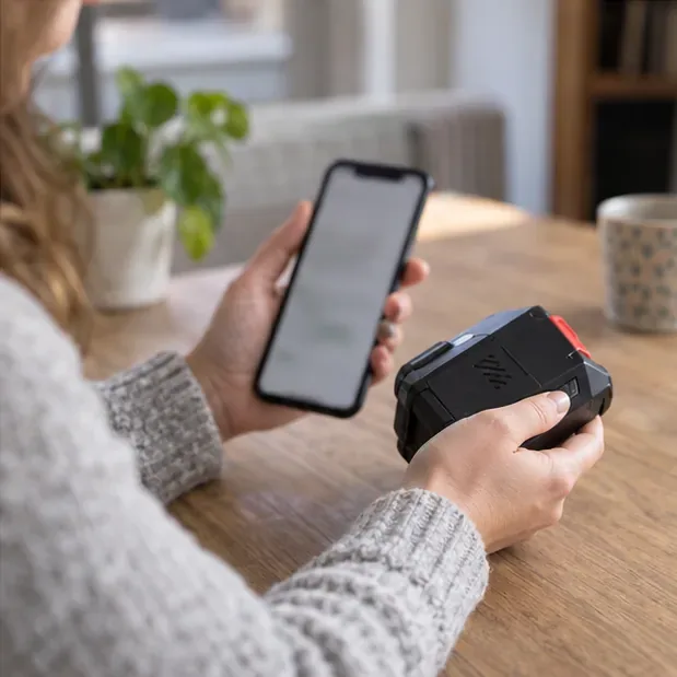 Een persoon bekijkt informatie op een smartphone terwijl een losse gereedschapsaccu op tafel ligt