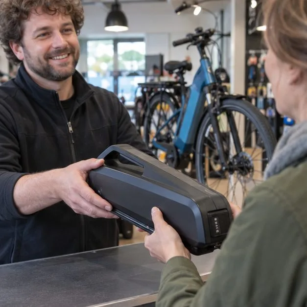 Een klant overhandigt een grote fietsaccu aan een medewerker bij een fietsenwinkel