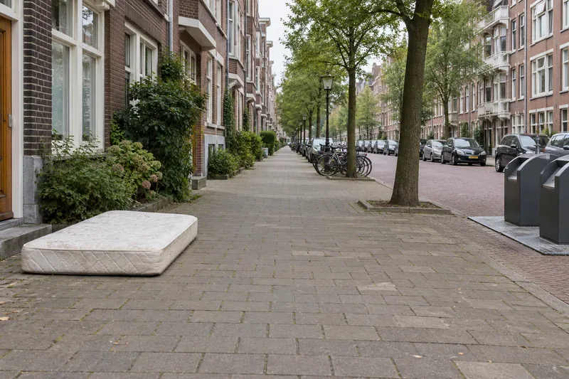 Een matras staat aan de zijkant van een brede stoep, op afstand van afvalcontainers en met vrije doorgang