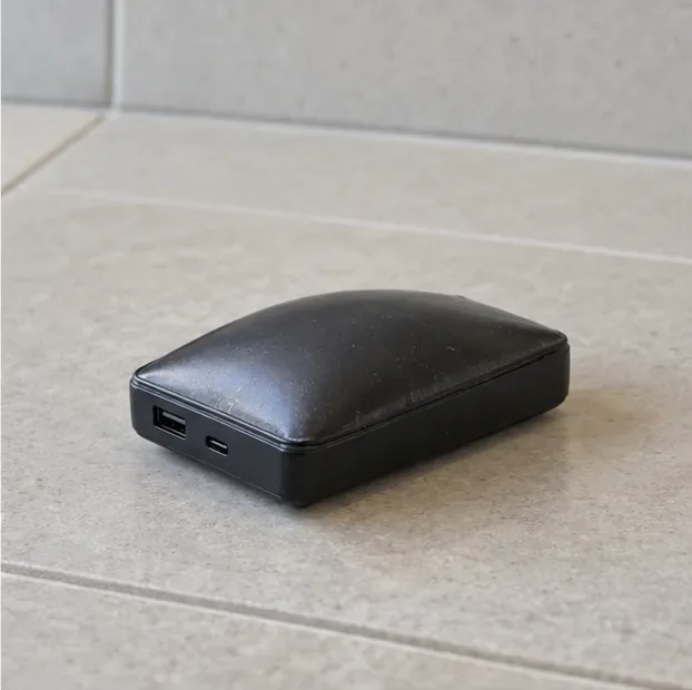 Een zwarte powerbank met een bolstaande bovenkant ligt alleen op een lichte, harde tegelvloer.