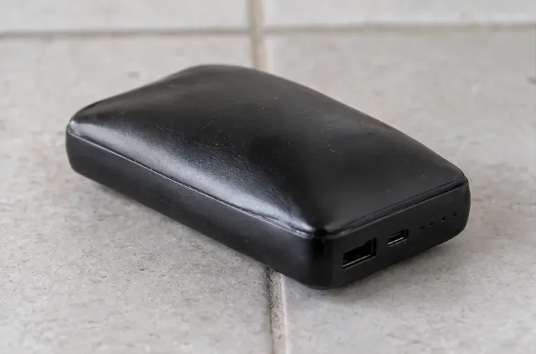 Een zichtbaar bolstaande zwarte powerbank ligt los op een keramische tegelvloer en is niet aangesloten