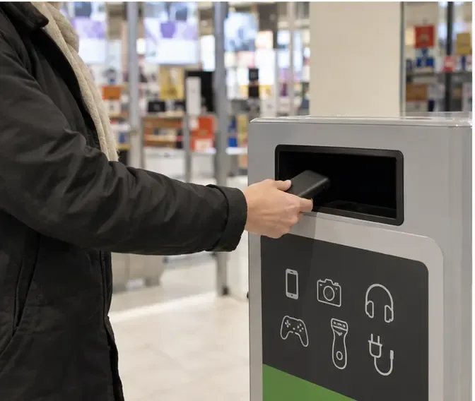 Een persoon plaatst een kleine zwarte powerbank in een inzamelmeubel voor kleine elektrische apparaten in een winkel