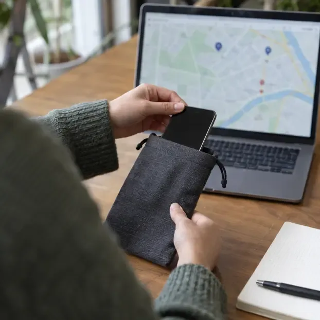 Een persoon schuift een uitgeschakelde smartphone in een beschermend zakje naast een laptop met een kaartweergave