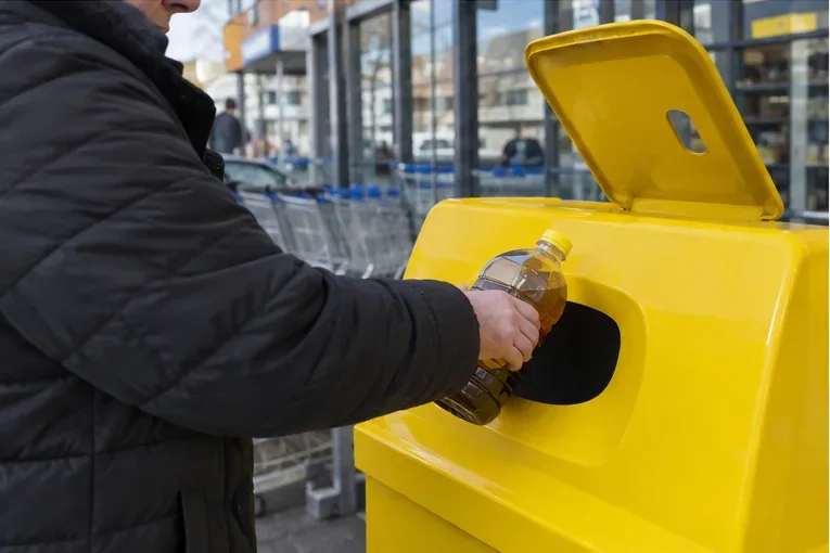 Persoon die een afgesloten fles met gebruikt frituurvet in een gele inzamelcontainer bij een winkel plaatst