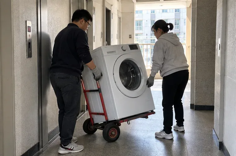두 사람이 운반용 손수레에 드럼세탁기를 올려 아파트 복도와 엘리베이터 쪽으로 이동하는 모습
