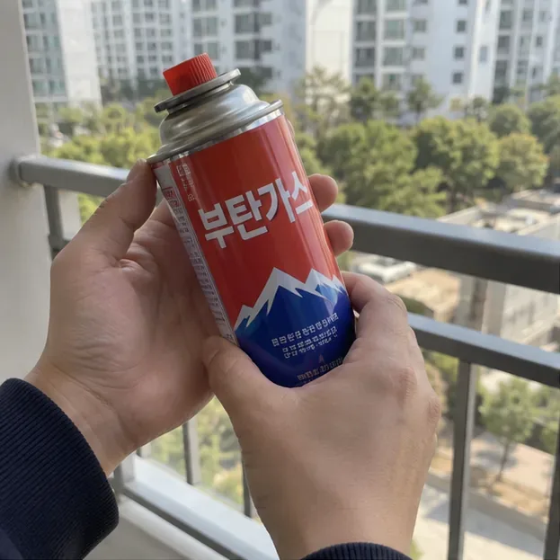 아파트 야외 공간에서 손에 든 휴대용 부탄가스통의 외관과 노즐 상태를 확인하는 모습