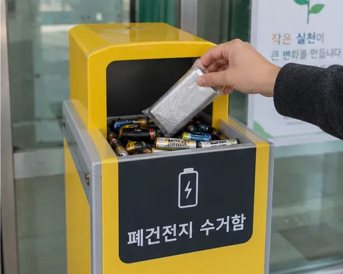 투명하게 포장한 보조배터리를 폐건전지 수거함에 넣는 손과 수거함 내부의 여러 건전지가 보이는 모습