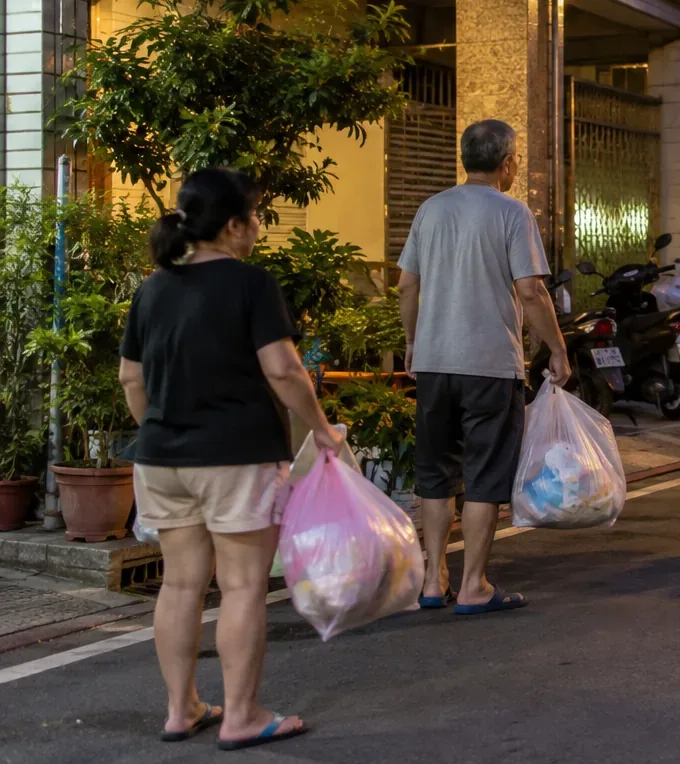兩位居民提著垃圾袋在住宅街道路邊步行，準備前往垃圾車清運位置。