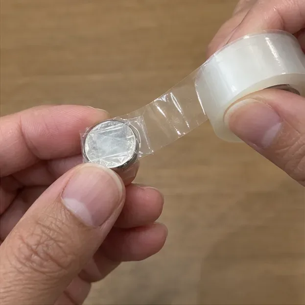 指で持った銀色のボタン電池の表面を透明テープで覆って絶縁している手元