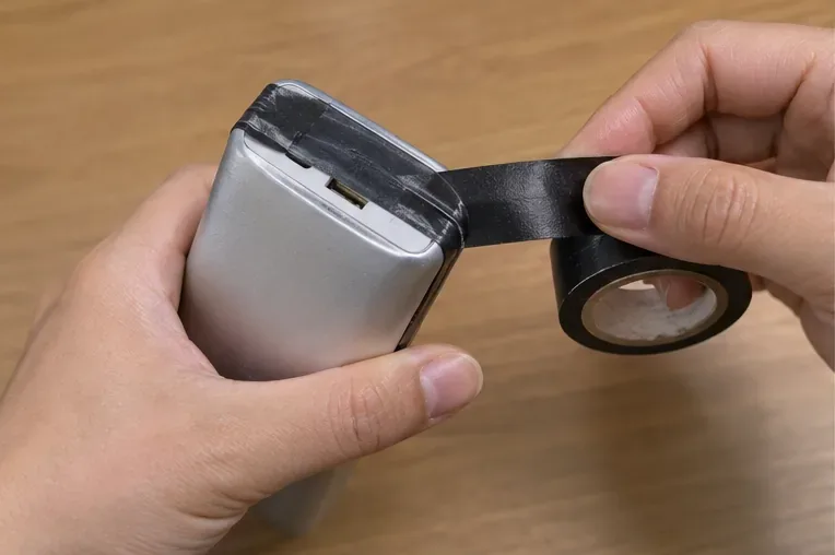 雙手在銀色行動電源連接埠附近貼上黑色絕緣膠帶，動作避免壓迫膨脹外殼