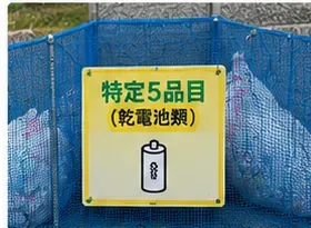 青いネット状のごみ集積場に「特定5品目（乾電池類）」と表示された案内板がある