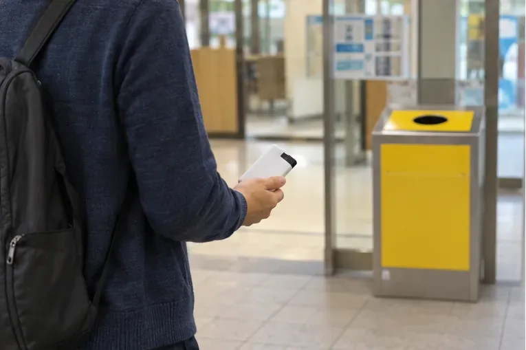 端子を絶縁したモバイルバッテリーを手に持ち、公共施設のロビーに設置された回収ボックスへ向かう様子