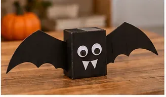 Piccola creatura pipistrello di cartone nero con ali attaccate, occhi rotondi e zanne bianche