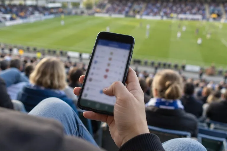 Aficionado en la grada consulta un teléfono móvil con el campo de fútbol desenfocado al fondo.