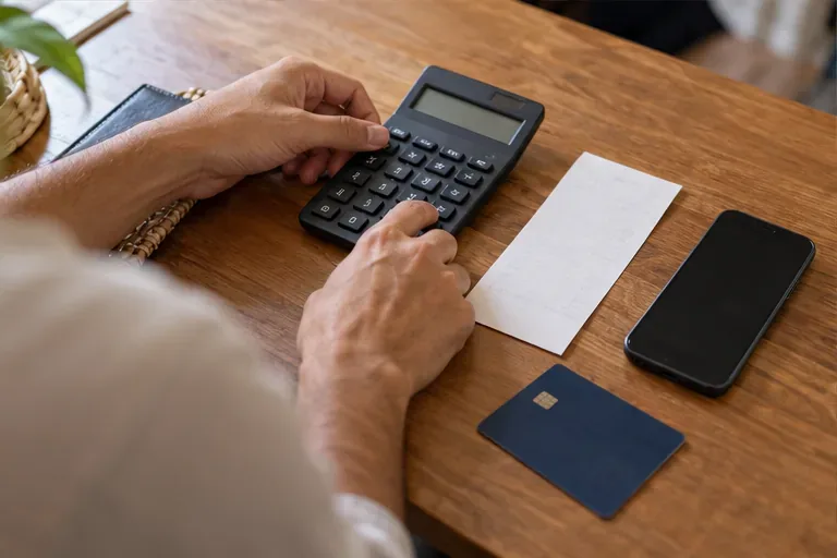 Mãos usam uma calculadora ao lado de um celular, cartão bancário e folha de papel.