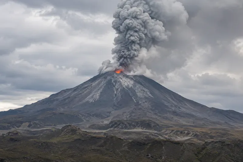 A gray ash plume rises from a steep volcanic cone, with a small orange glow near the summit and rocky ground in the foreground