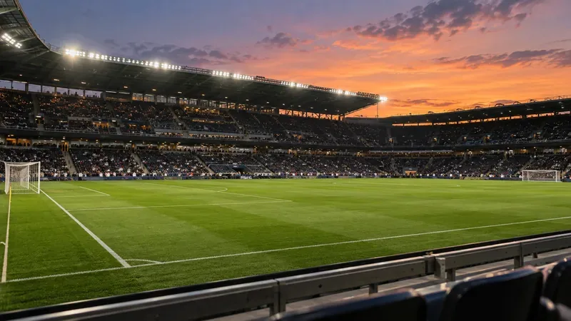Een groen voetbalveld met witte lijnen en twee doelen, voor een stadion met tribunes bij avondlicht.