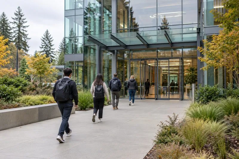 Employees walk toward a modern glass technology office building on an overcast morning