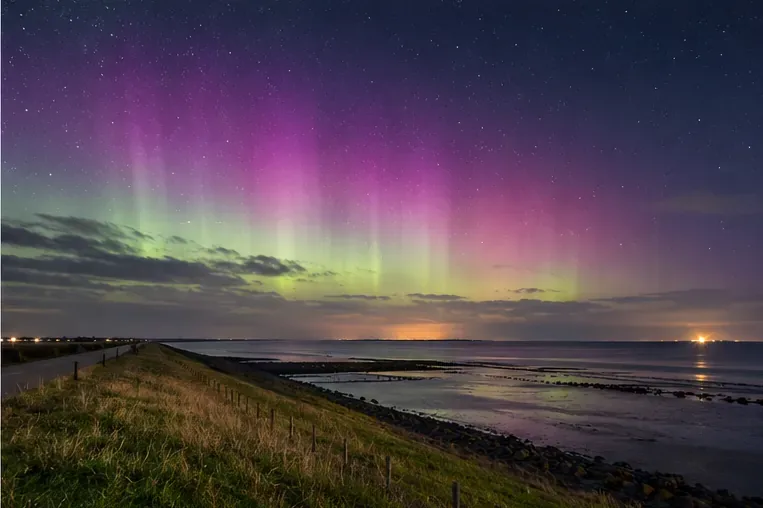 Brede kustdijk en ondiepe Waddenzee onder roze en groene noorderlichtsluiers