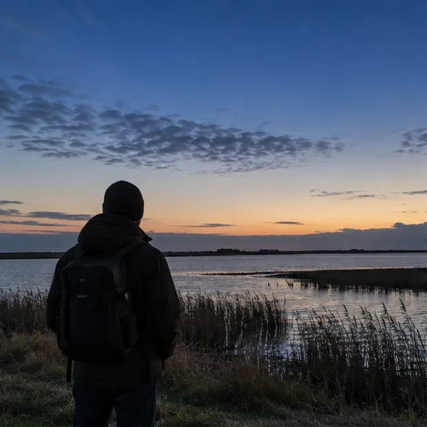 Persoon met rugzak kijkt over riet en water bij schemering, met vrije horizon
