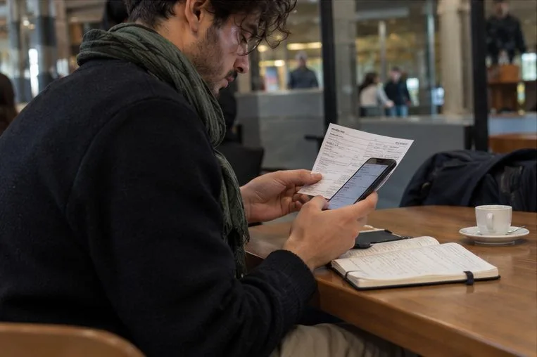 Un passeggero confronta il telefono, il biglietto e gli appunti a un tavolo vicino alla stazione.