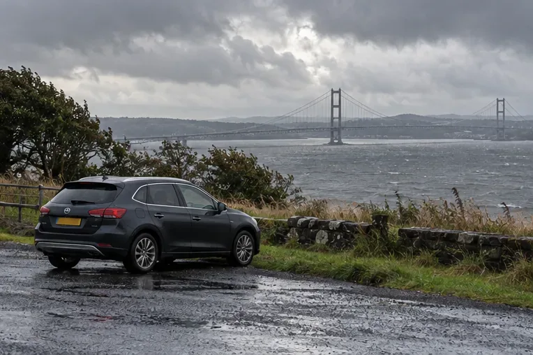 A car waits off the roadway beside a windy estuary, with a suspension bridge in the distance.