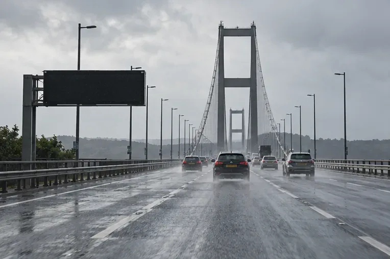 Cars approach a suspension bridge on a wet road beneath a blank overhead variable-message sign.