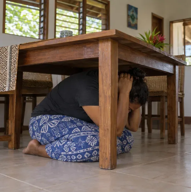 A person crouches under a sturdy wooden table and shields their head during an earthquake drill.